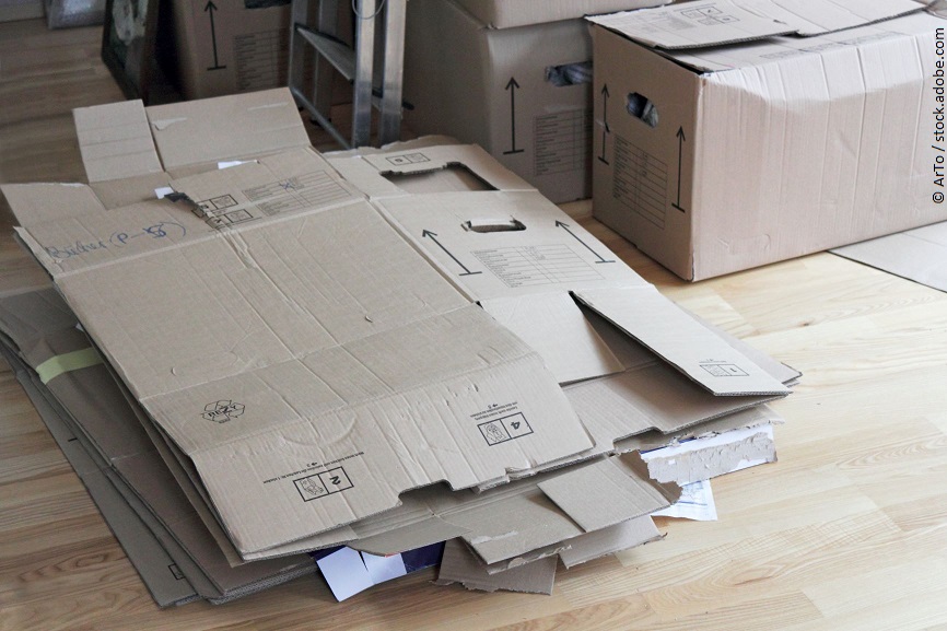 Zusammengefaltene Umzugskarton liegen auf einem Laminatboden. Dahinter stehen volle Umzugskartons. 